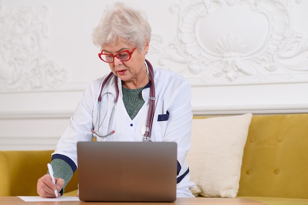 Ärztin mittleren alters, die laptop-notizen mit stethoskop auf dem tisch schreibt, gesundheitskonzept med