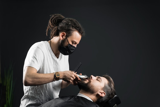 Afeitado de bigote en peluquería. Barbero con rastas en máscara médica negra recorta la barba de un hombre guapo en la cuarentena coronavirus covid-19.