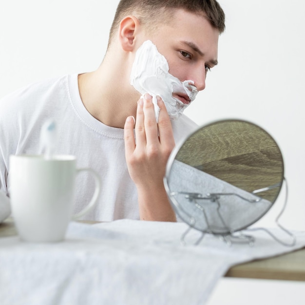Afeitado diario Higiene facial Rutina de cuidado de la piel Concentrado joven apuesto mirando en el espejo aplicando espuma sobre la barba en el baño blanco