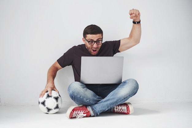 Aficionado al fútbol emocionado con una pelota de fútbol aislado sobre fondo blanco. Se sienta en el suelo y observa los resultados del partido y los mejores momentos del partido en el portátil.