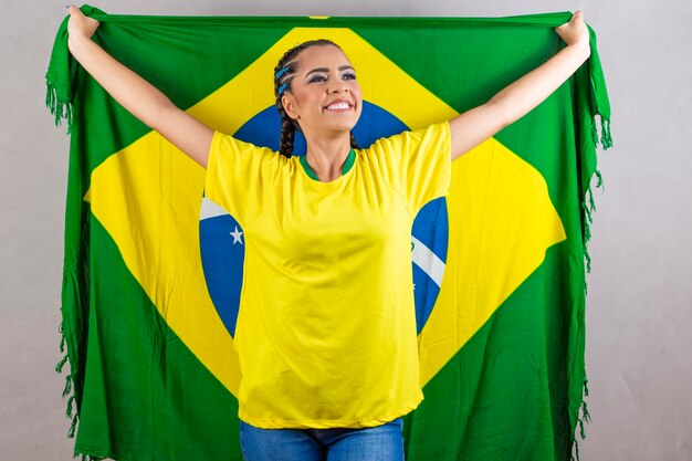 Aficionado brasileño sosteniendo la bandera de Brasil sobre fondo gris Joven mujer brasileña sonriente sonriendo a la cámara con la bandera de Brasil