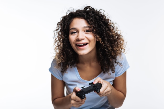 Aficionado a los juegos. Hermosa mujer alegre de pelo rizado jugando videojuegos con un controlador de juego y sonriendo en gris