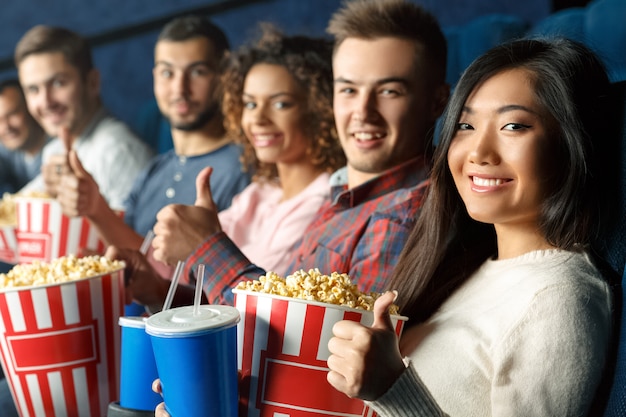 Aficionados al cine todos aquí. Grupo de amigos alegres que muestran los pulgares juntos sentados en el cine