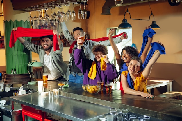 Aficionados al deporte animando en el bar, pub y bebiendo cerveza mientras se lleva a cabo el campeonato, la competencia. Grupo multiétnico de amigos emocionado viendo la traducción. Las emociones humanas, la expresión, el concepto de apoyo.