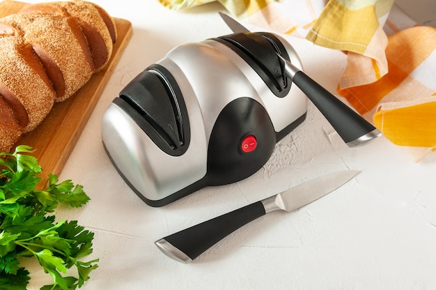 Afilador de cuchillos eléctrico. el cuerpo de plástico es gris-negro. un  cuchillo de cocina en el afilador. en la parte de atrás hay pan y verduras.  fondo claro.