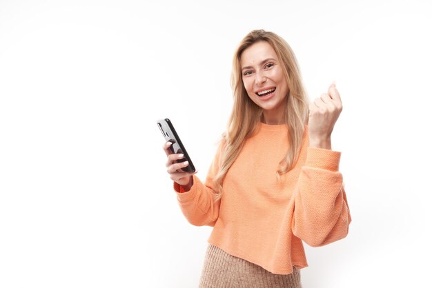 Afortunada chica rubia sonriente sosteniendo el teléfono inteligente en casual aislado en el fondo blanco del estudio recibió un mensaje alegre, celebrando la victoria