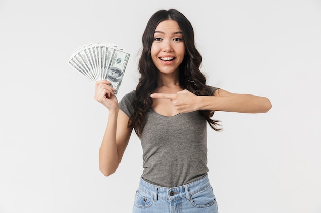 Afortunada mujer morena de 20 años vestida con ropa básica sonriendo y sosteniendo un montón de billetes de dinero aislados sobre la pared blanca