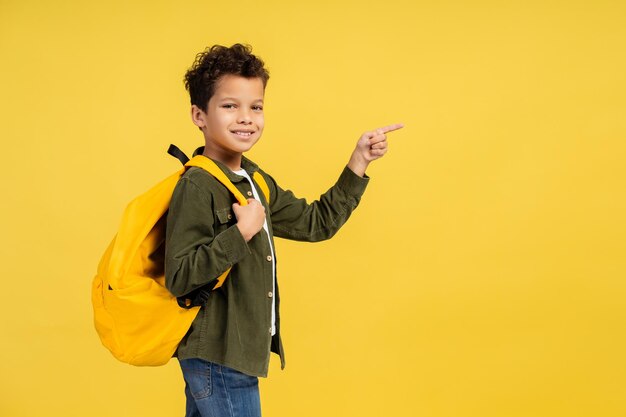 Foto african american boy with yellow backpack pointing to copy space isolated on yellow background