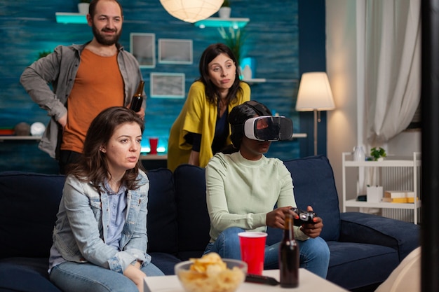 Africana experimentando la realidad virtual jugando videojuegos mientras sus amigos socializan. Grupo de personas de raza mixta pasando el rato juntos divirtiéndose a altas horas de la noche en la sala de estar.