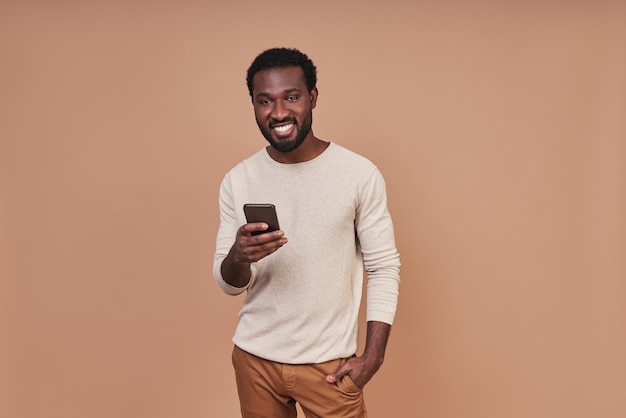 Africano jovem bonito em roupas casuais, usando um telefone inteligente e sorrindo