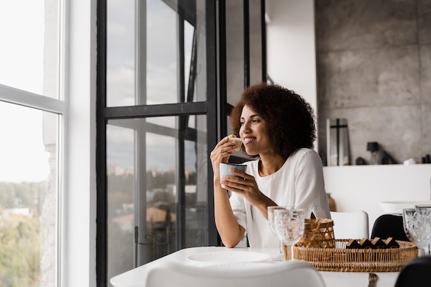 Afrikanerin, die Cupcake isst und Kaffee trinkt und sich während einer Pause von der Arbeit in der Wohnung entspannt Afrikanisch-amerikanisches Mädchen isst im Hotel am Esstisch in der Nähe von großen Fenstern zu Mittag