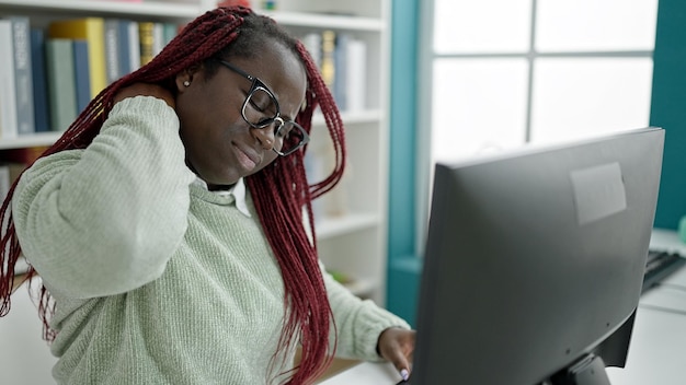 Foto afrikanerin mit geflochtenem haar, studentin benutzt computer mit nackenschmerzen in der universitätsbibliothek