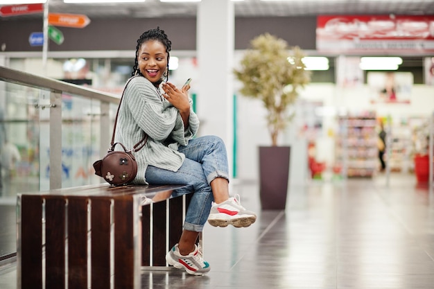 Afrikanerin posierte in Pullover und Jeans und posierte in einem Einkaufszentrum, das mit ihrem Handy auf einer Bank saß