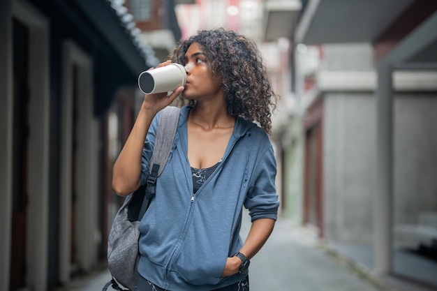 Afrikanerin sport gesund fit und fest schlank trinkt Wasser aus der Tasse auf der Hand Fitness-Mädchen trägt Sportbekleidung und Jacke zu Fuß in der Stadt und in der Stadt Fitness- und Sportmotivation