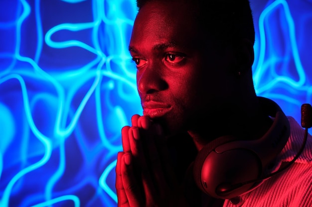 Afrikanisch-amerikanischer Mann mit Headset am Hals, der mit gefalteten Händen gegen bunte leuchtende Neonlichter denkt