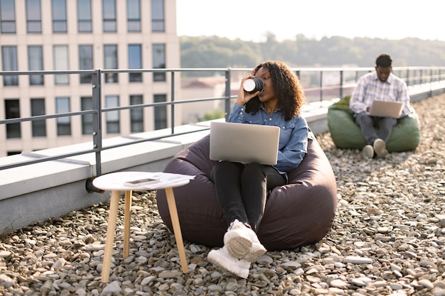 Afrikanische Dame hält eine Tasse Getränk, während sie Webseiten auf einem tragbaren Computer durchläuft