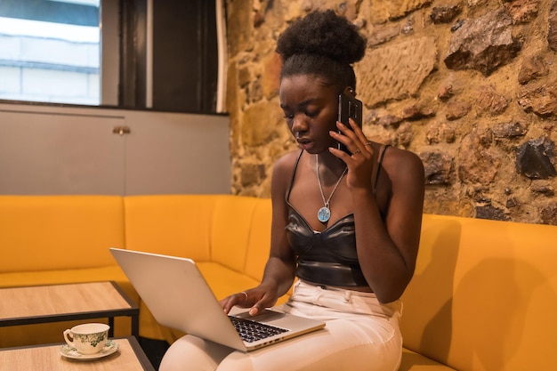 Afrikanische Frau arbeitet mit einem Laptop und spricht mit dem Handy in einer Cafeteria