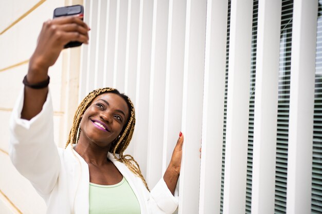 Afrikanische Frau, die ein Selfie mit einem Handy mit einem glücklichen Gesichtsausdruck macht, der an eine Wand gelehnt ist