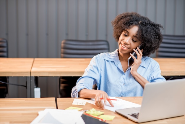 Afrikanische Frau, die mit dem Telefon spricht, das mit Laptop und Dokumenten im modernen Bürointerieur sitzt