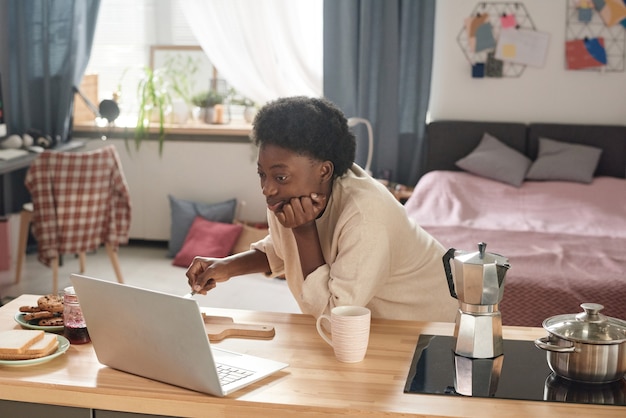 Afrikanische Frau, die morgens während ihres Frühstücks etwas auf dem Laptop am Tisch beobachtet