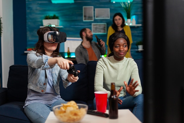 Afrikanische Frau, die während des Videospielwettbewerbs für Freunde jubelt und eine Virtual-Reality-Brille trägt, die auf dem Sofa sitzt und Spaß hat und einen drahtlosen Controller verwendet. Gemischte Rassengruppe von Menschen, die Geselligkeit haben.