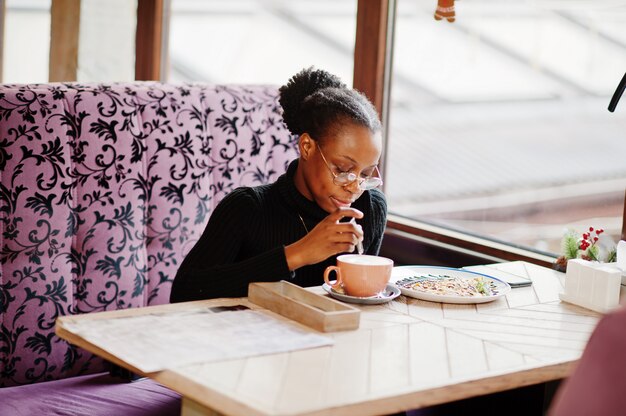 Afrikanische Frau im schwarzen Pullover und in den Brillen stellte im Café auf, sitzend am Tisch mit Nachtisch und Tasse Kaffee.