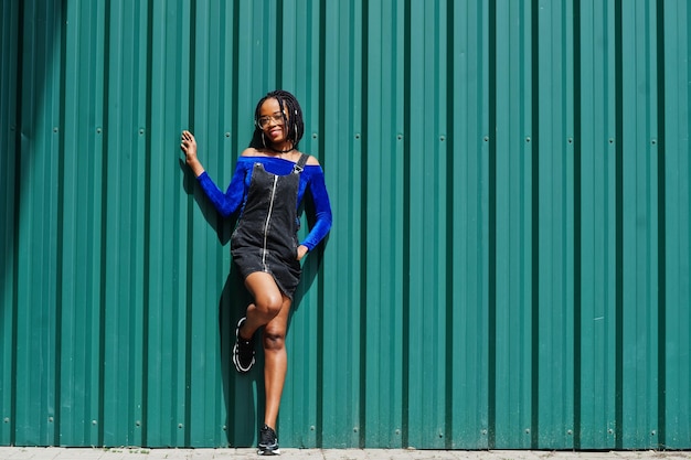 Afrikanische Frau in Denim-Overalls und Rockbrillen posierte vor grüner Stahlwand