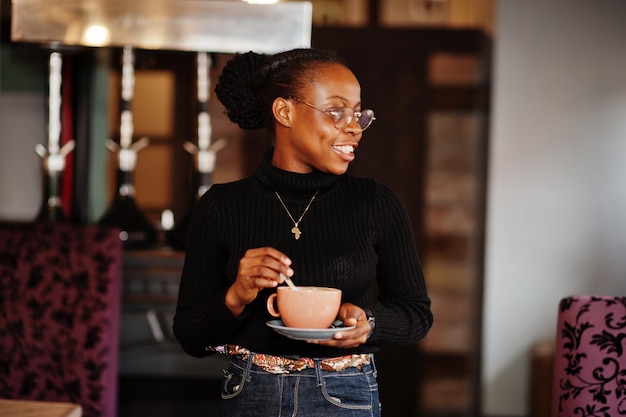 Afrikanische Frau in schwarzem Pullover und Brille posierte im Café mit einer Tasse heißem Getränk