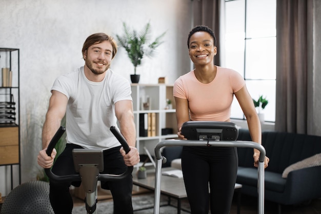 Afrikanische Frau läuft auf Laufband und kaukasischer Mann trainiert auf Heimtrainer