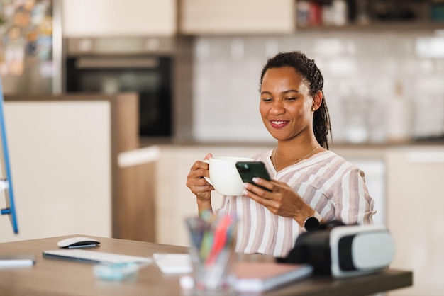 Afrikanische Geschäftsfrau benutzt ein Mobiltelefon und trinkt Kaffee, während sie von zu Hause aus arbeitet.