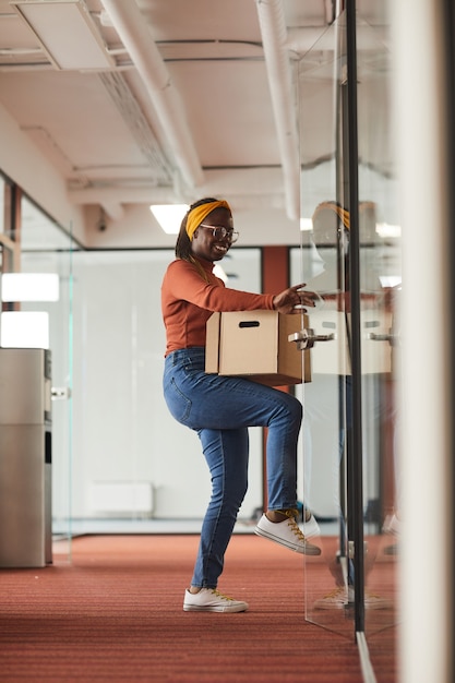 Afrikanische Geschäftsfrau in Freizeitkleidung, die Kiste mit Dingen hält und die Tür öffnet, die sie im Büro ist