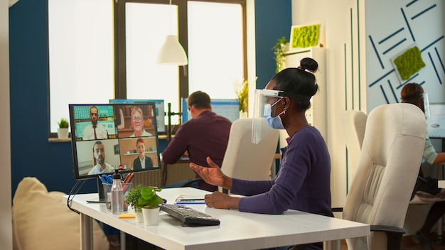 Afrikanische Geschäftsfrau mit Gesichtsmaske im Büro, die während der Coronavirus-Epidemie per Videoanruf mit dem Remote-Team spricht. Online-Konferenz, Videomeeting-Kollegen, die unter Wahrung der sozialen Distanz arbeiten.