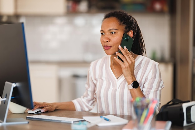 Afrikanische Geschäftsfrau spricht auf einem Smartphone, während sie von ihrem Heimbüro aus am Computer arbeitet.
