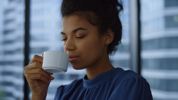 Afrikanische Geschäftsfrau trinkt Kaffee im modernen Büro Porträt nachdenklicher Manager blickt in Bürofenster Geschäftsfrau genießt Kaffeetasse im Büro mit Blick auf die Stadt Nachdenklicher Berufstätiger in der Pause