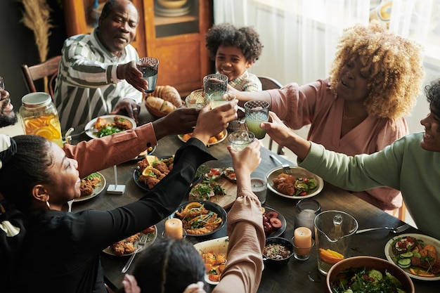 Afrikanische Großfamilie, die mit Gläsern mit Limonade röstet, während sie gemeinsam am Tisch zu Abend essen