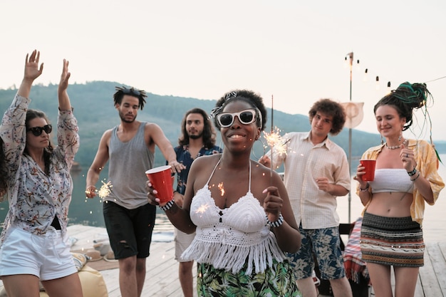 Afrikanische junge Frau in Sonnenbrille mit Glas Bier und Wunderkerzen, die an der Kamera lächeln, während sie mit ihren Freunden an der Party im Freien tanzen