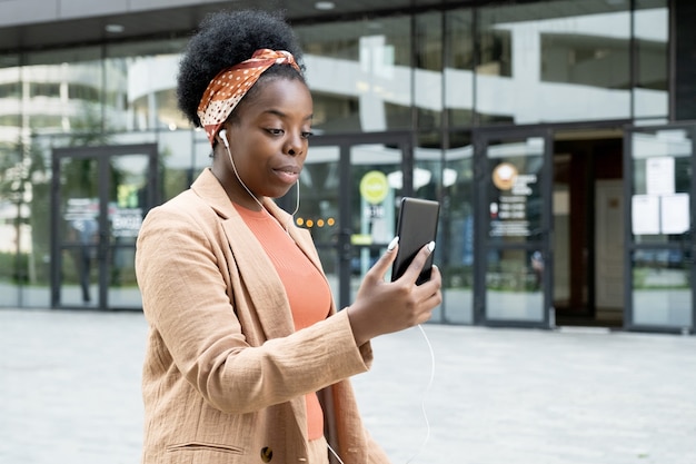 Afrikanische junge Frau mit Kopfhörern, die online mit ihrem Handy spricht, während sie die Stadtstraße entlang geht