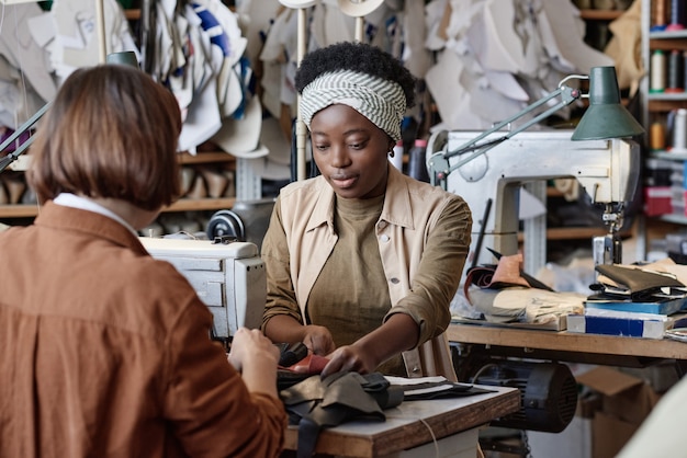 Afrikanische junge Schneiderin, die zusammen mit ihrer Kollegin Kleidung aus Leder näht, während sie am Tisch sitzen und an der Nähmaschine arbeiten