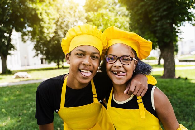 Afrikanische Kinderköche in Kochmütze und gelben Uniformen, die Grimassen schneiden und sich gegenseitig die Zunge zeigen Afrikanischer Teenager und schwarzes Mädchen haben Spaß und kochen Essen