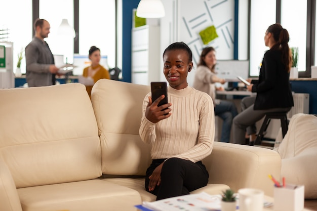 Afrikanische Managerin, die mit entfernten Kollegen über Videoanrufe diskutiert, die Smartphone mit Kopfhörern halten, die auf der Couch im modernen Büro des Geschäfts sitzen