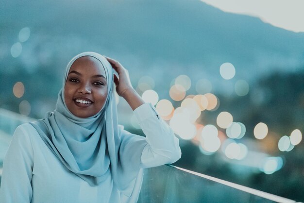 Afrikanische muslimische Frau in der Nacht auf einem Balkon, der in die Kamera lächelt, mit Bokeh-Lichtern der Stadt im Hintergrund. Hochwertiges Foto