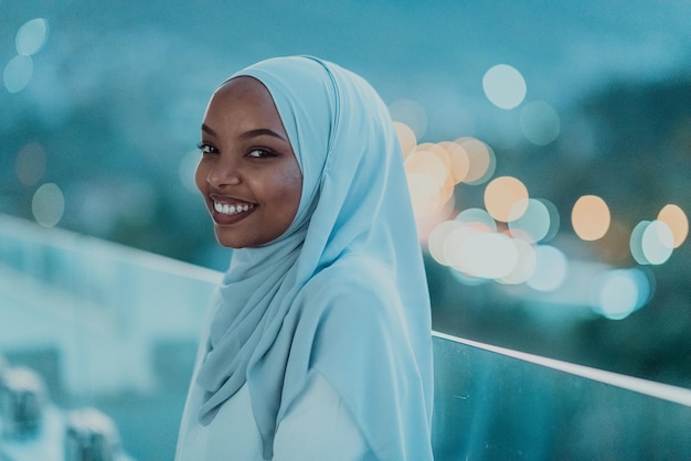 Afrikanische muslimische Frau in der Nacht auf einem Balkon, der in die Kamera lächelt, mit Bokeh-Lichtern der Stadt im Hintergrund. Hochwertiges Foto