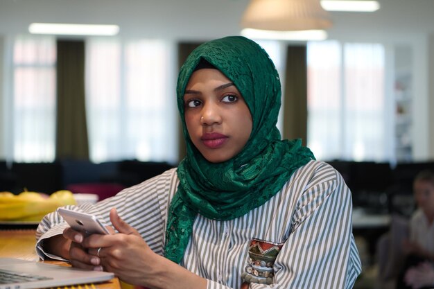 afrikanische muslimische geschäftsfrau, die einen grünen hijab trägt und ein handy benutzt, während sie an einem laptop im entspannungsbereich eines modernen großraum-startbüros arbeitet. Vielfalt, gemischtrassiges Konzept