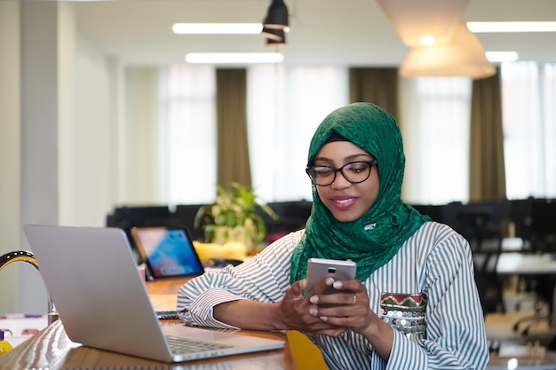 afrikanische muslimische geschäftsfrau, die einen grünen hijab trägt und ein handy benutzt, während sie an einem laptop im entspannungsbereich eines modernen großraum-startbüros arbeitet. Vielfalt, gemischtrassiges Konzept