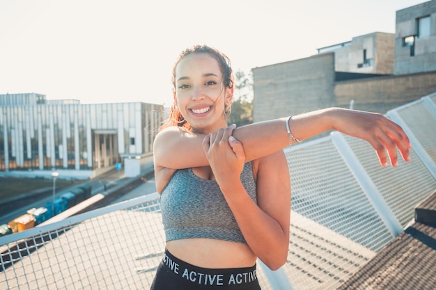 Afrikanische Sportfrau, die ihre Arme ausdehnt, während sie zum städtischen Sportszenario der Kamera lächelt. Sonnenuntergang. Schönheit gesunde arabische Frau. Sportkleidung, neue Gewohnheiten für ein neues Jahr, abnehmen.