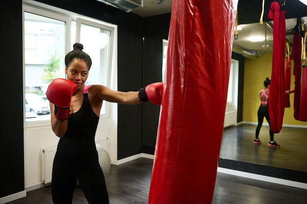Afrikanische Sportlerin, Boxerin mit perfektem Körperbau mit roten Boxhandschuhen, auffallend auf Boxsack im Sportstudio, mit Spiegelung im Spiegel. Kampfkunst des Kampfes. Gesunder aktiver Lebensstil