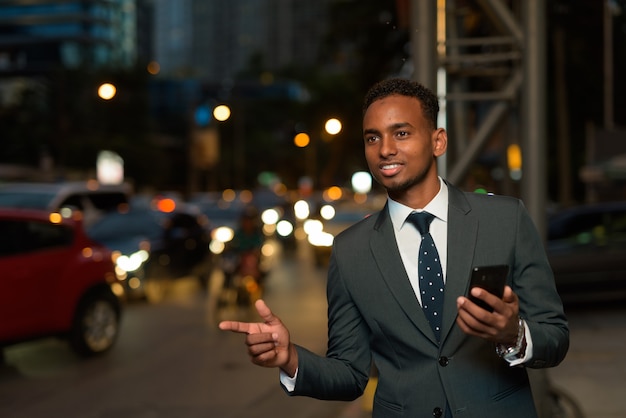 Afrikanischer Geschäftsmann, der Handy-App verwendet, die auf Taxi mit Hand oben in der Nacht wartet