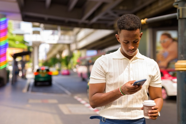 Afrikanischer Geschäftsmann, der legere Kleidung trägt, während er das Handy benutzt und eine Kaffeetasse am Taxistand in der Stadt hält?