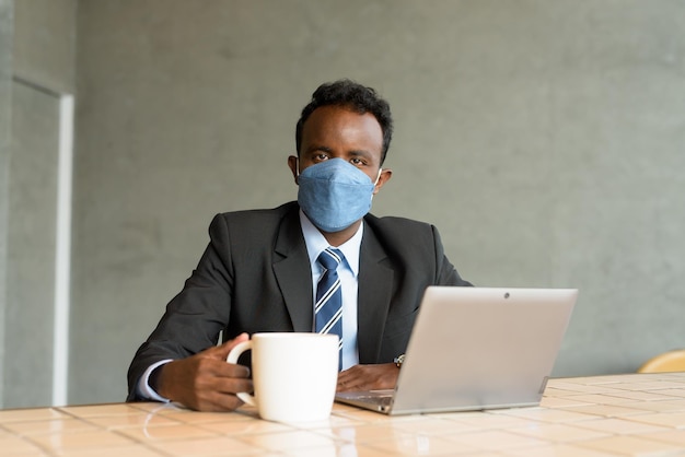 Afrikanischer Geschäftsmann im Café, während er einen Laptop benutzt und eine Gesichtsmaske trägt, während er sich sozial distanziert