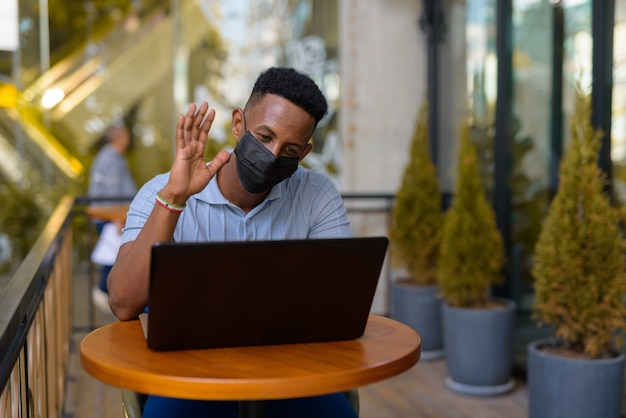 Afrikanischer Geschäftsmann mit Gesichtsmaske und sozialer Distanzierung beim Sitzen im Café mit Laptop-Computer und Videoanruf beim Lächeln